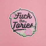 Circular, pastel pink vinyl sticker with the words Fuck the Tories in a black script. The sticker is decorated with pink roses, green foliage and white sparkles, all bursting over the edge of the circle. The sticker has a white edge.