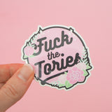 Circular, pastel pink vinyl sticker with the words Fuck the Tories in a black script. The sticker is decorated with pink roses, green foliage and white sparkles, all bursting over the edge of the circle. The sticker has a white edge and is held between a finger and thumb.