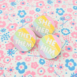 Three round enamel pins on a pastel, floral background. Each pin has pastel rainbow diagonal stripes. There is a short horizontal line in the centre with pronouns above and below. They read individually in white capitals; She/Her, He/Him, or They/Them.
