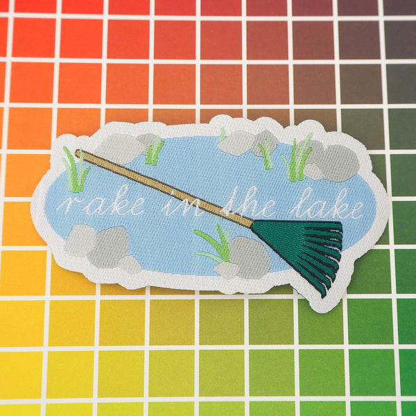 Woven patch showing an oval lake with stones and weeds around its edge. Across the lake is a large green rake and the words Rake in the Lake in white script. The patch has a white border.