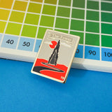 Tall rectangular enamel pin showing a spiked black tower on a red plain. A winding black road leads to the tower. A red sun and whispy clouds hang in a grey sky behind the tower. A grey ribbon banner at the top of the pin reads The Tower in silver letters. A small, silver number 19 is written in the top left and bottom right corner. The pin is shown resting against a colourful notepad with a numbered grid.