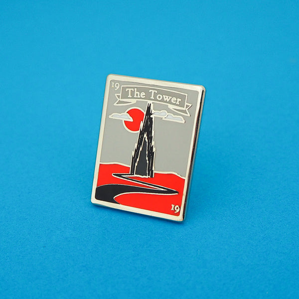 Tall rectangular enamel pin showing a spiked black tower on a red plain. A winding black road leads to the tower. A red sun and whispy clouds hang in a grey sky behind the tower. A grey ribbon banner at the top of the pin reads The Tower in silver letters. A small, silver number 19 is written in the top left and bottom right corner. The pin is shown on a blue background.