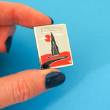 Tall rectangular enamel pin showing a spiked black tower on a red plain. A winding black road leads to the tower. A red sun and whispy clouds hang in a grey sky behind the tower. A grey ribbon banner at the top of the pin reads The Tower in silver letters. A small, silver number 19 is written in the top left and bottom right corner. The pin is being held between a forefinger and thumb.
