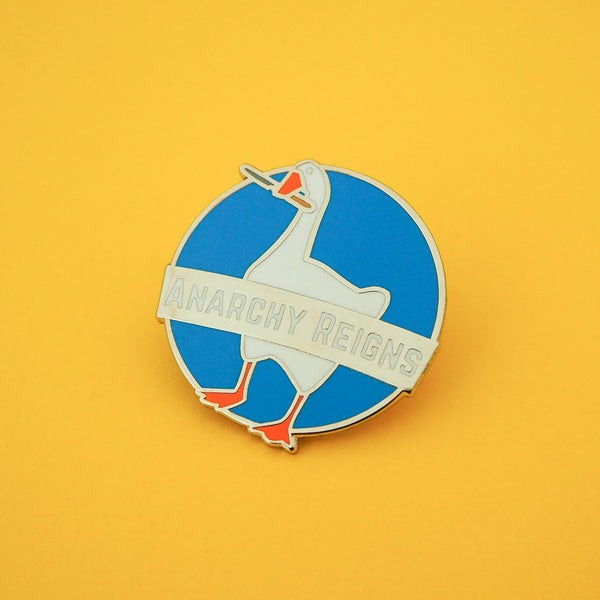 A circular, blue enamel pin depicting a goose with a knife in its beak. A banner across the middle reads Anarchy Reigns. The pin is shown on a bright yellow background.