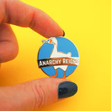 A circular, blue enamel pin depicting a goose with a knife in its beak. A banner across the middle reads Anarchy Reigns. The pin is being held between a forefinger and thumb above a bright yellow background.