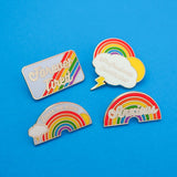 Four enamel pins. The first is a rectangle reading Forever Tired on diagonal rainbow stripes.  The second reads Professional Overthinker on a lightning cloud with sun and rainbow. The third has a rainbow with sun and lightning cloud and reads Leave Me Alone. The fourth depicts a rainbow with the word Anxious in white script.
