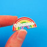 An enamel pin depicting a rainbow with the word Anxious in white script. The pin is being held between a thumb and forefinger over a blue background.