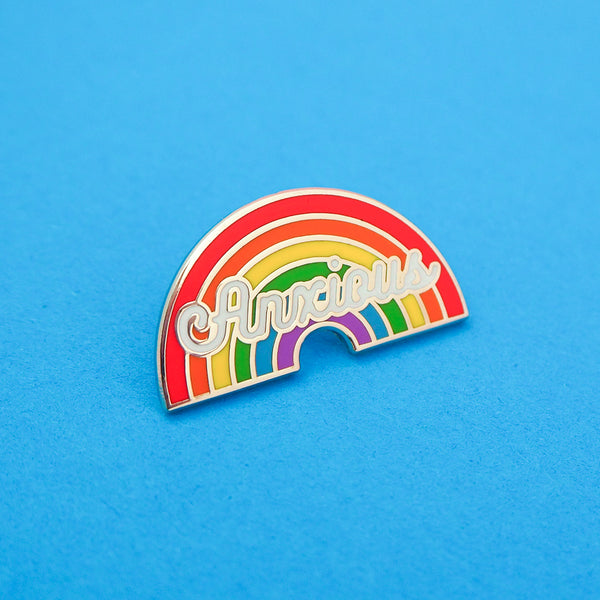 An enamel pin depicting a rainbow with the word Anxious in white script. The pin is on a blue background.