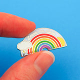 Metal pin showing a rainbow with sun and lightning cloud that reads Leave Me Alone in silver script. The pin is held between finger and thumb.