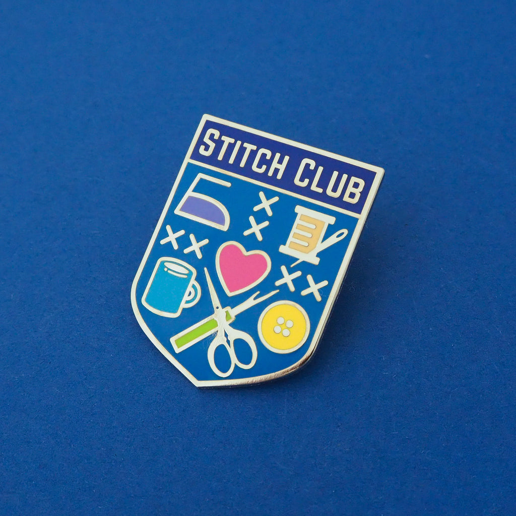 Bright blue, shield-shaped enamel pin with the words Stitch Club at the top in capital letters in a dark blue banner. The heart of the shield contains many images of brightly coloured sewing paraphernalia including a button, needle and thread, scissors and seam-ripper. Pin is shown on a deep blue background.