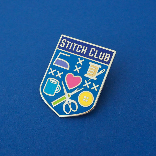 Bright blue, shield-shaped enamel pin with the words Stitch Club at the top in capital letters in a dark blue banner.  The heart of the shield contains many images of brightly coloured sewing paraphernalia including a button, needle and thread, scissors and seam-ripper. Pin is shown on a deep blue background.