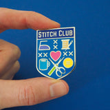 Bright blue, shield-shaped enamel pin with the words Stitch Club at the top in capital letters in a dark blue banner. The heart of the shield contains many images of brightly coloured sewing paraphernalia including a button, needle and thread, scissors and seam-ripper. The pin is held between a thumb and forefinger.
