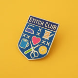 Bright blue, shield-shaped enamel pin with the words Stitch Club at the top in capital letters in a dark blue banner. The heart of the shield contains many images of brightly coloured sewing paraphernalia including a button, needle and thread, scissors and seam-ripper. Pin is shown on a yellow background.