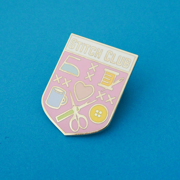 Pastel pink, shield-shaped enamel pin with the words Stitch Club at the top in capital letters in a white banner. The heart of the shield contains many images of brightly coloured sewing paraphernalia including a button, needle and thread, scissors and seam-ripper. Pin is shown on a blue background.