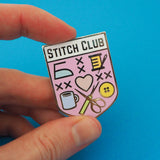 Pastel pink, shield-shaped enamel pin with the words Stitch Club at the top in capital letters in a white banner. The heart of the shield contains many images of brightly coloured sewing paraphernalia including a button, needle and thread, scissors and seam-ripper. Pin is being held between a forefinger and thumb.