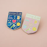 Two enamel pins on a pastel pink background. One is a bright blue, shield-shaped pin with the words Stitch Club at the top in capital letters in a dark blue banner. The heart of the shield contains many images of brightly coloured sewing paraphernalia including a button, needle and thread, scissors and seam-ripper. The second is the same design but the background is pastel pink with a white banner.