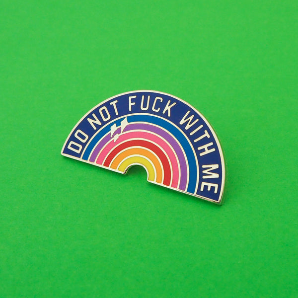 Rainbow enamel pin that reads Do Not Fuck With Me in silver capital letters on a dark blue arch. The colourful rainbow beneath has three silver sparkles in the upper left. The pin is shown on a bright green background.