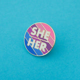 Round enamel pin with blue, purple, and pink diagonal stripes. There is a short horizontal line in the centre; above it is the word She and below it, Her. The lettering is white block capitals. The pin is on a light blue background.