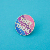 Round enamel pin with blue, purple, and pink diagonal stripes. There is a short horizontal line in the centre; above it is the word They and below it, Them. The lettering is white block capitals. The pin is on a light blue background.