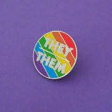 Round enamel pin with bright rainbow diagonal stripes. There is a short horizontal line in the centre; above it is the word They and below it, Them. The lettering is white block capitals. The pin is on a deep purple background.