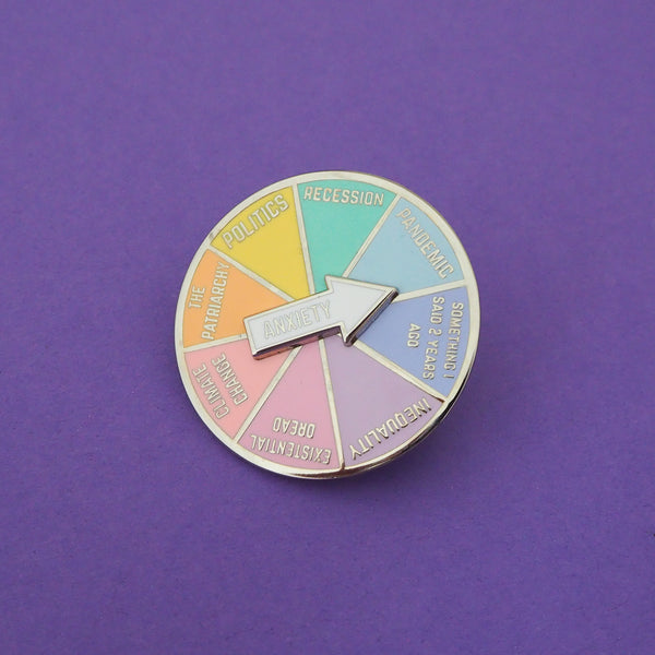 A circular pin with a spinning arrow which reads Anxiety. The main body of the pin is split into 8 pastel-coloured sections. They are labelled; Recession, Pandemic, Something I Said 2 Years Ago, Inequality, Existential Dread, Climate Change, The Patriarchy, Politics.