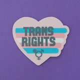Heart-shaped pastel pink woven patch with horizontal stripes in the colours of the trans flag. The words Trans Rights are written in grey capitals on the stripes. The stripes extend slightly beyond the heart on the right with rounded edges. At the bottom of the heart is a grey transgender symbol. The patch has a pastel pink border.