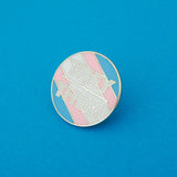 Round enamel pin with blue, pink, and white diagonal stripes. There is a short horizontal line in the centre; above it is the word They and below it, Them. The lettering is white block capitals. The pin is on a bright blue background.