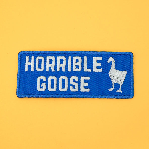 Rectangular, blue embroidered patch that reads Horrible Goose in white capitals with a goose silhouette in white on the right. The patch has a blue merrowed edge.