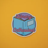 Vinyl sticker showing an open book with a turquoise cover. The words Library Enthusiast are written in pink script. The sticker has a white border.