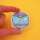 Vinyl sticker showing an open book with a turquoise cover. The words Library Enthusiast are written in pink script. The sticker has a white border and is held between a finger and thumb.