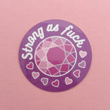 Round, purple vinyl sticker. It has a multi-faceted pink gem in the centre. At the top are the words Strong as fuck and below the gem are small hearts in shades of pink.