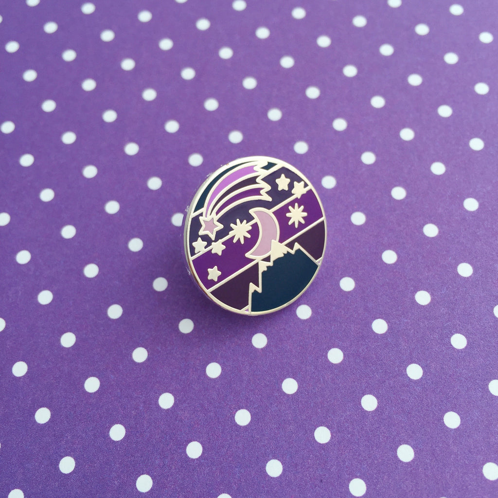 Round metal pin showing a moon and stars over a mountain on a striped background. Above the moon there is a shooting star with a long tail. The colours are a variety of dark blues and purples.