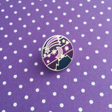Round metal pin showing a moon and stars over a mountain on a striped background. Above the moon there is a shooting star with a long tail. The colours are a variety of dark blues and purples.