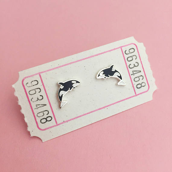 A pair of enamel stud earrings depicting orca whales mid-jump. One is facing right and the other faces left. The earrings are on a white carnival ticket.