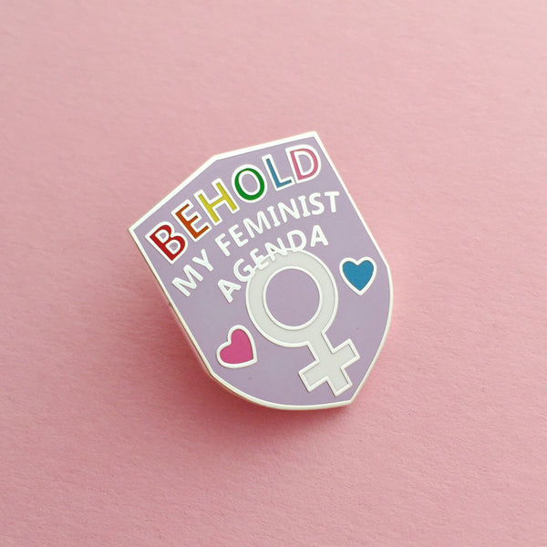 Pink enamel pin in a shield shape. At the top it reads Behold My Feminist Agenda. The word Behold is filled in rainbow colours. At the bottom there is a feminine symbol with a pink heart to the left and a blue heart to the right. The pin is on a pink background.