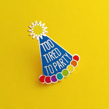 Enamel pin depicting a blue conical party hat. The hat has rainbow bobbles at the base and a yellow pom-pom on top. The hat reads Too Tired To Party in silver capitals.