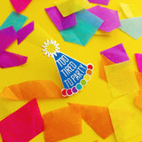 Enamel pin depicting a blue conical party hat. The hat has rainbow bobbles at the base and a yellow pom-pom on top. The hat reads Too Tired To Party in silver capitals. The pin is amongst brightly coloured confetti.