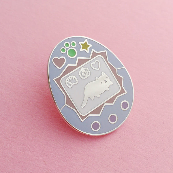 A light blue enamel pin showing an egg-shaped virtual pet toy. On the white screen is a silver cat and game icons. Beneath the screen are 3 purple buttons and above are a heart, pawprint and star in pastel colours.