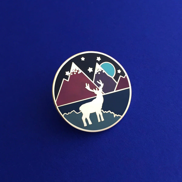 Round enamel pin showing a silver silhouette of a stag standing on a lakeshore in front of mountains. The colours are rich, deep blues, greens and purples. There is a full moon and stars in the sky.
