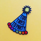 Embroidered patch depicting a blue conical party hat. The hat has rainbow bobbles at the base and a yellow pom-pom on top. The hat reads Too Tired To Party in black capitals. The patch has black merrowed edges.