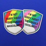 Two shield-shaped vinyl stickers that read I'm Not Sorry About Your Fragile Masculinity in black capitals. One sticker has a bright rainbow in the background and the other has a pastel rainbow. The stickers have a white border.