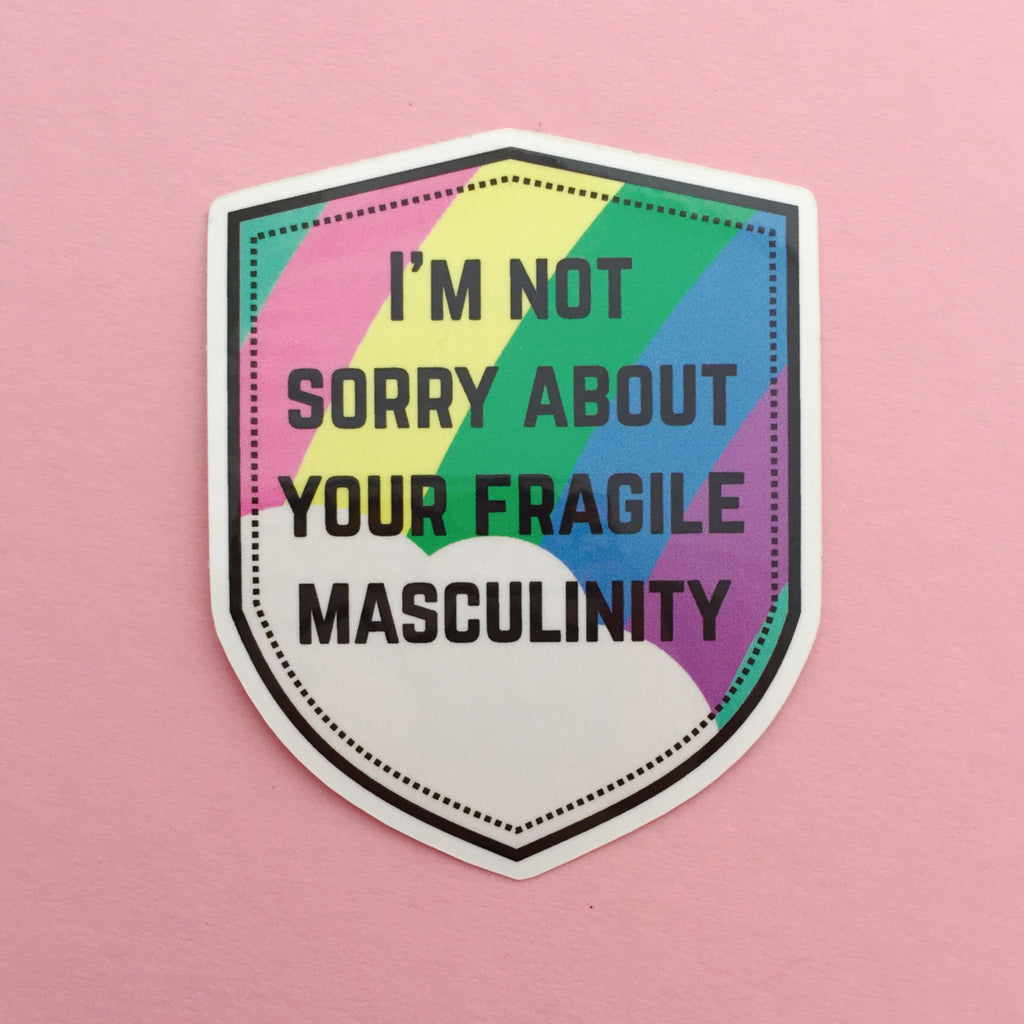 Shield-shaped vinyl sticker that reads I'm Not Sorry About Your Fragile Masculinity in black capitals. Behind the words is a pastel rainbow and cloud. The sticker has a white border.