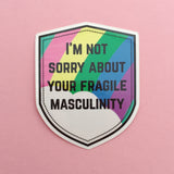 Shield-shaped vinyl sticker that reads I'm Not Sorry About Your Fragile Masculinity in black capitals. Behind the words is a pastel rainbow and cloud. The sticker has a white border.