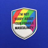 Shield-shaped vinyl sticker that reads I'm Not Sorry About Your Fragile Masculinity in black capitals. Behind the words is a bright rainbow and cloud. The sticker has a white border.