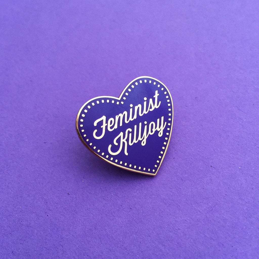 A deep purple enamel pin in the shape of a heart. The words Feminist Killjoy are written in rose gold in a flowing script. Rose gold dots run along the inside border of the heart. The pin is shown on a purple background.
