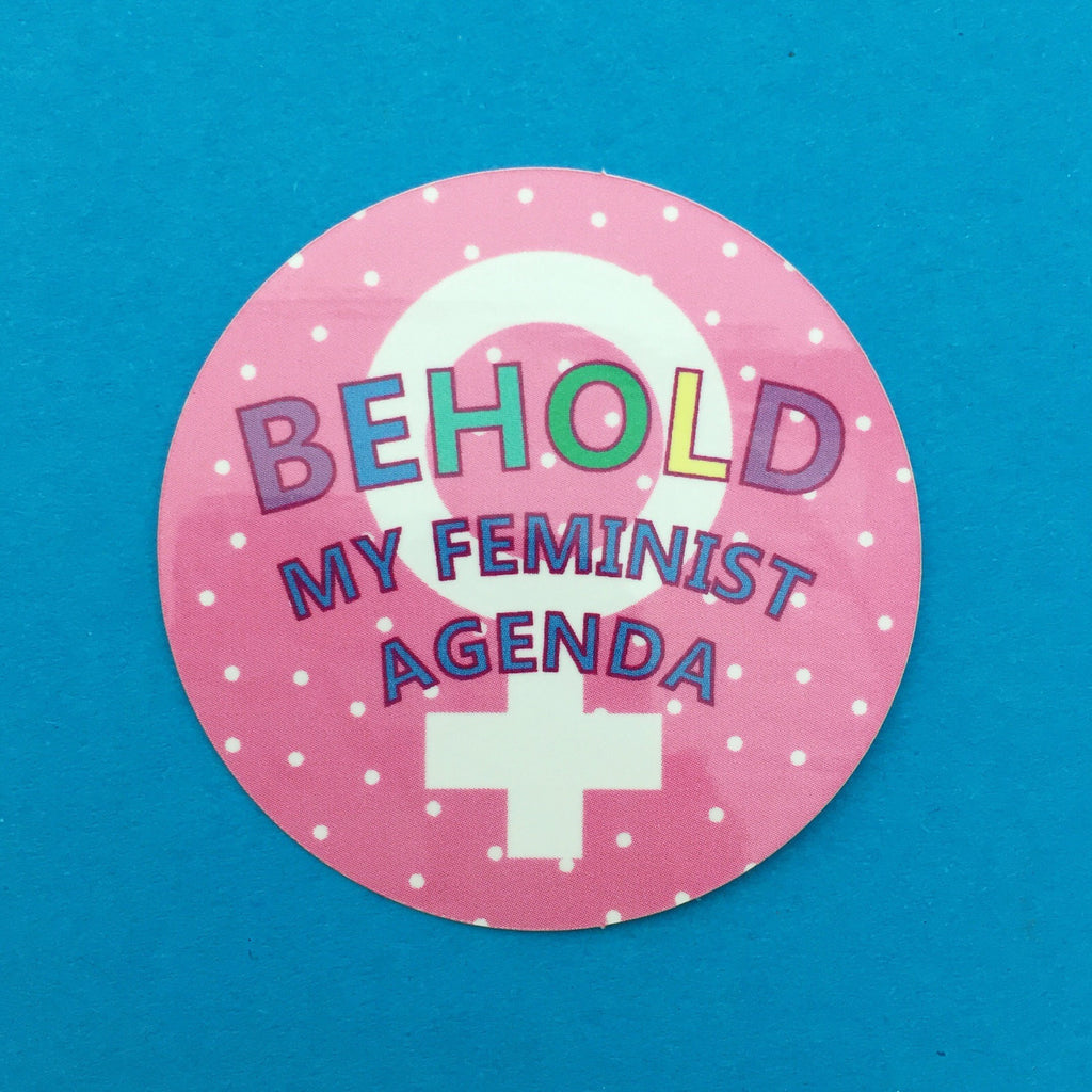 Round, pink sticker. It reads Behold My Feminist Agenda. The word Behold is filled in rainbow colours. Behind the words are a white feminine symbol and white polka-dots.