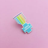 Enamel pin shaped like a medal. The bar is purple and the ribbon section has pastel-rainbow vertical stripes. The medal is a pastel blue, rounded cross bearing the words Coping Admirably in capital letters. The pin is on a light pink background.