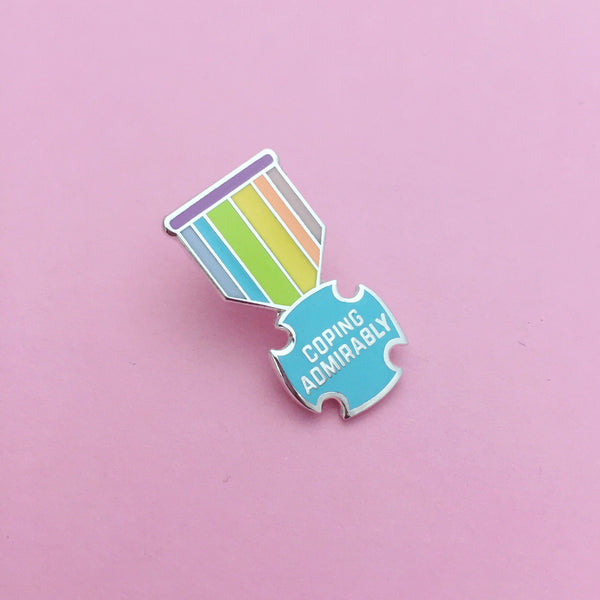 Enamel pin shaped like a medal. The bar is purple and the ribbon section has pastel-rainbow vertical stripes. The medal is a pastel blue, rounded cross bearing the words Coping Admirably in capital letters. The pin is on a light pink background.