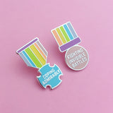 Two enamel pins shaped like a medals. On the first the bar is purple and the ribbon section has pastel-rainbow vertical stripes. The medal is a pastel blue, rounded cross bearing the words Coping Admirably in capital letters. The second has the pastel-rainbow ribbon above the purple bar, which is thicker. The medal is pastel pink, round, and bears the words Fighting Invisible Battles in capital letters. The pins are on a light pink background.