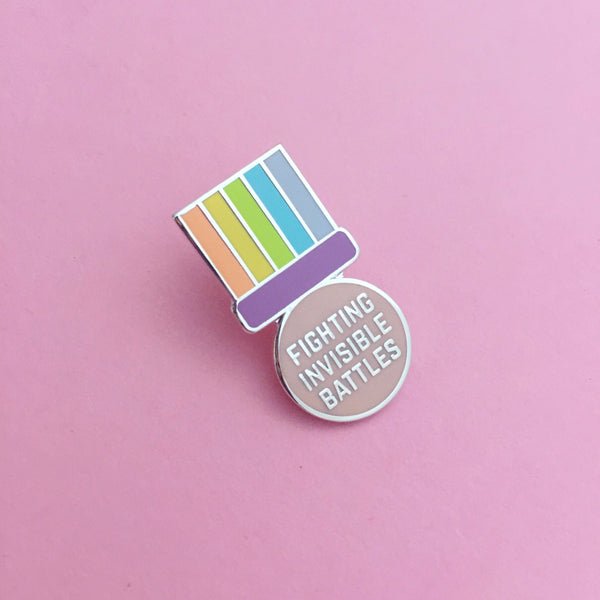 Enamel pin shaped like a medal. A pastel-rainbow ribbon section is above a purple bar. Below the bar, the medal is pastel pink, round, and bears the words Fighting Invisible Battles in capital letters. The pin is on a light pink background.